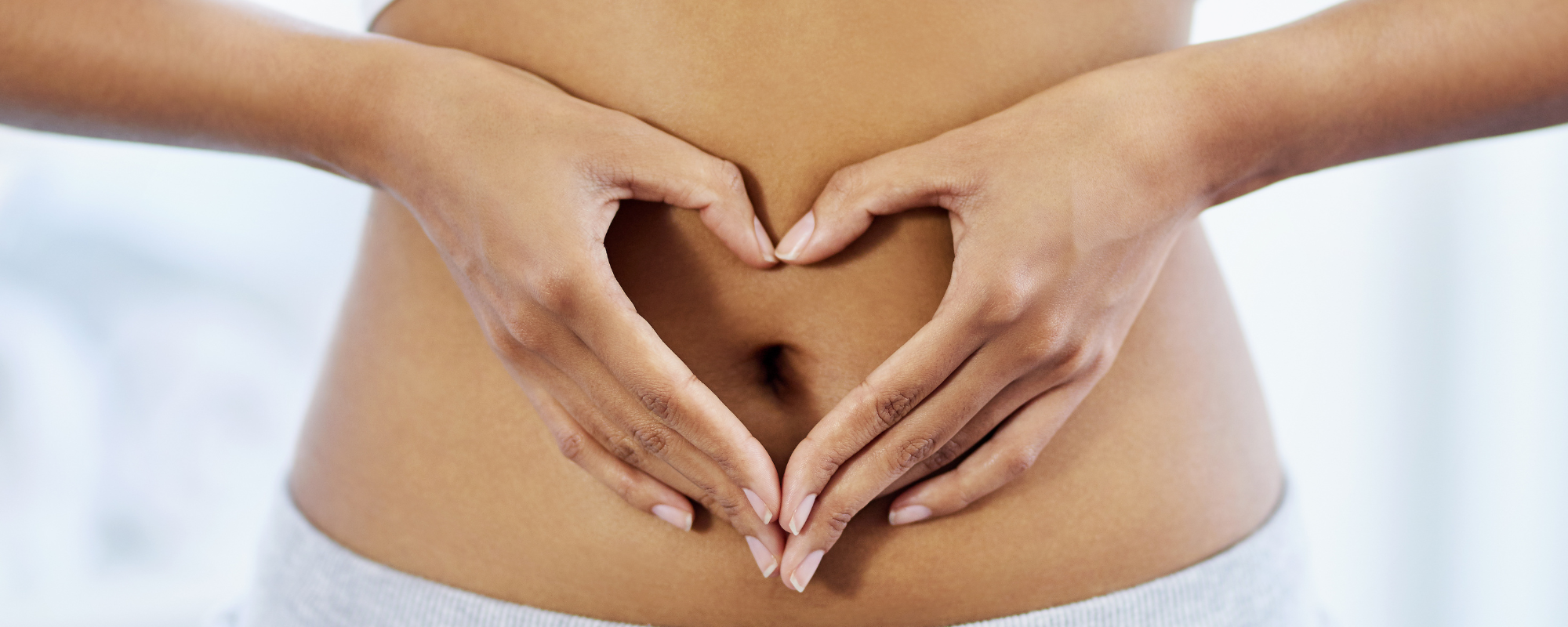 Close-up of hands forming a heart shape over a bare stomach.