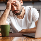 Tired man in white shirt resting head on hand next to a green mug and laptop at a table.