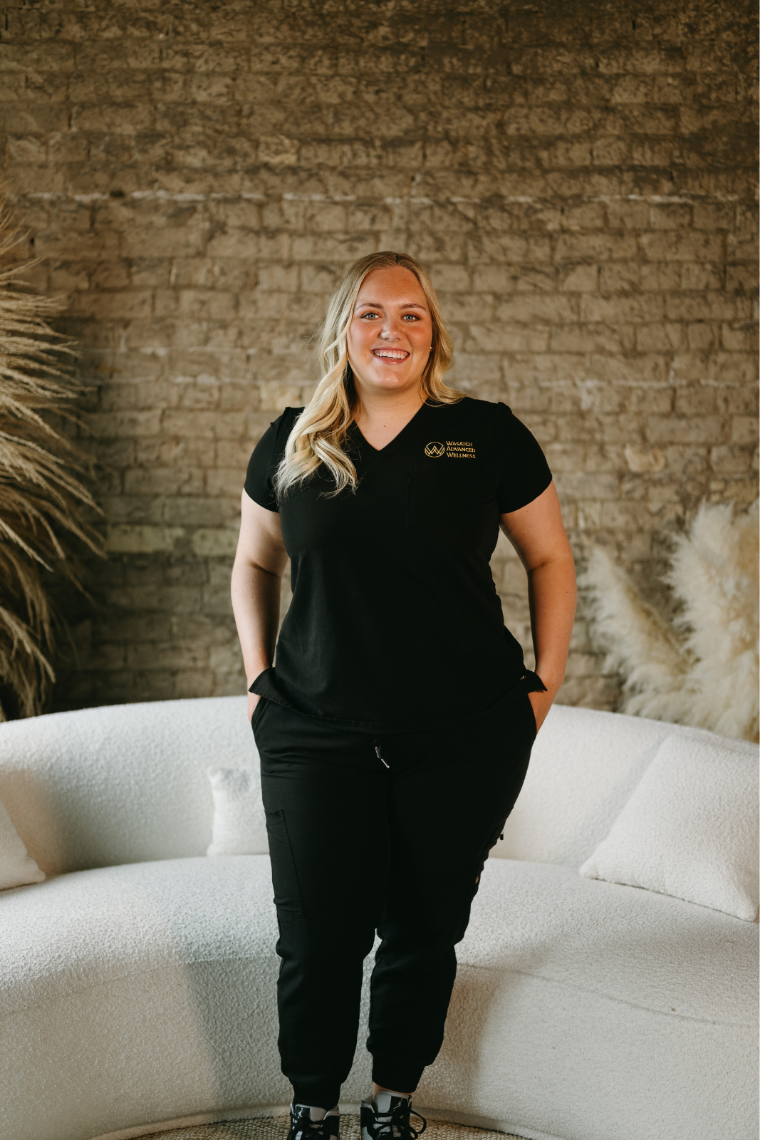 Chelsa Bringhurst NP Smiling woman with long wavy hair in black outfit and white coat sitting on textured couch.
