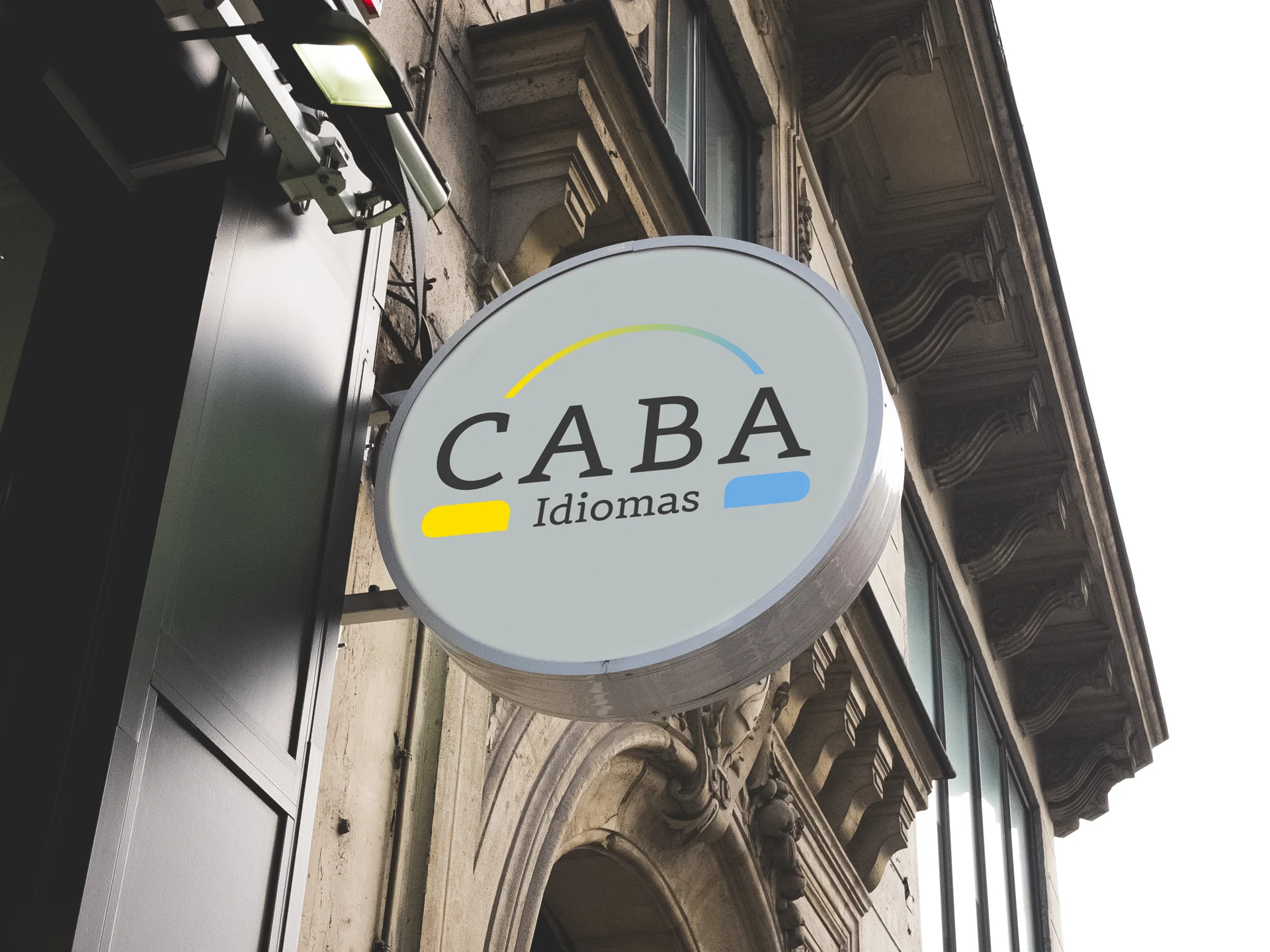 Placa circular da CABA Idiomas aplicada à fachada de um edifício, com identidade em amarelo, azul e cinza.