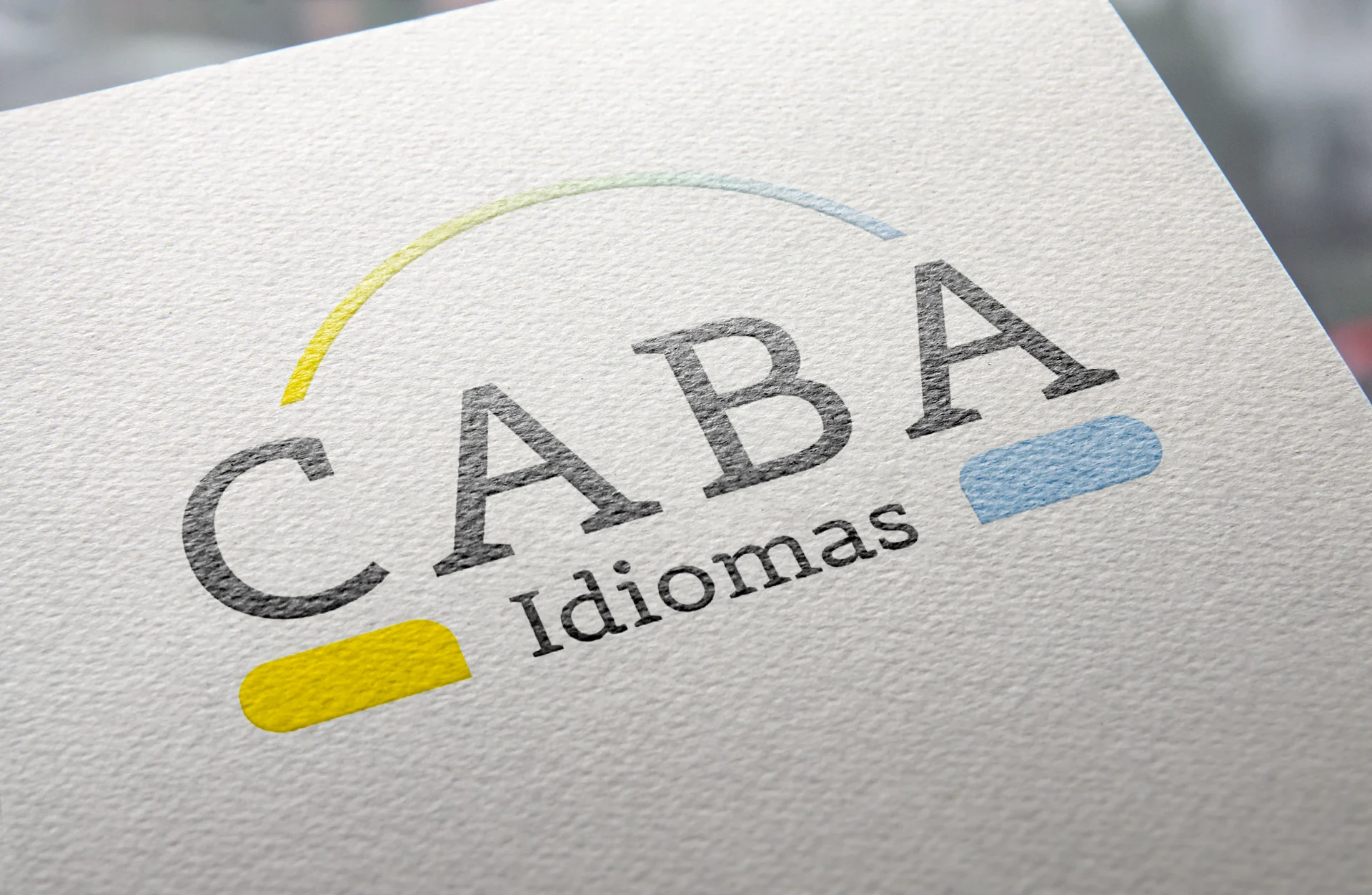 Detalhe do logotipo CABA Idiomas aplicado em papel texturizado, com arco em gradiente amarelo e azul.
