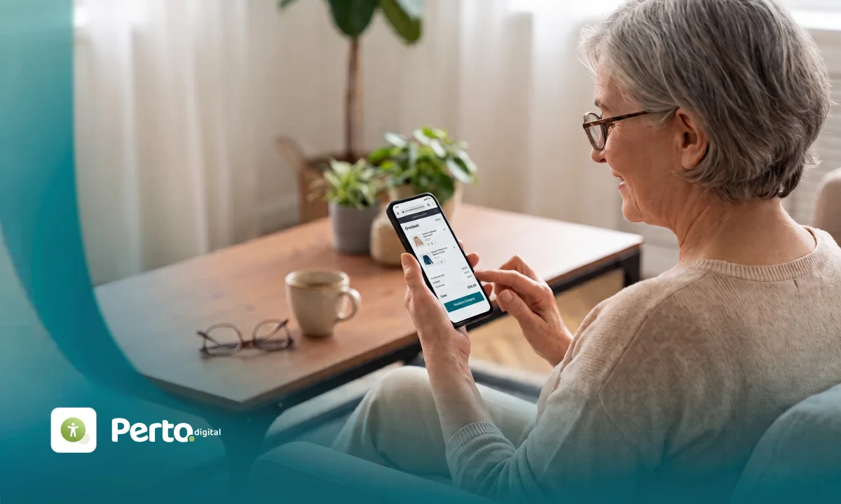 Mulher idosa de óculos e cabelos grisalhos vista de perfil, sentada no sofá de casa, enquanto segura um celular e finaliza uma compra online. No canto inferior esquerdo, está o logotipo da Perto Digital.
