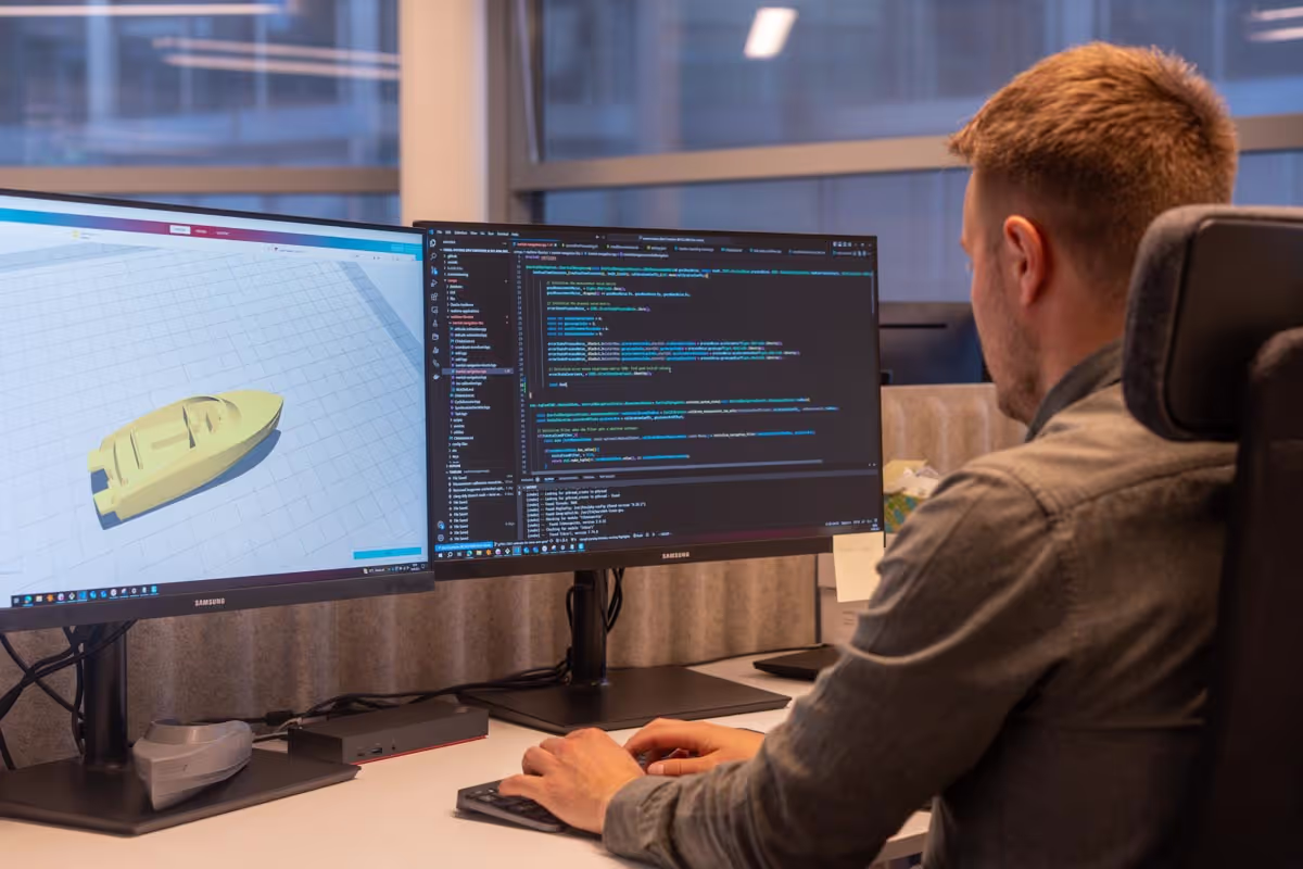 Man working on dual monitors showing 3D modeling on left screen and code editor on right screen in an office.