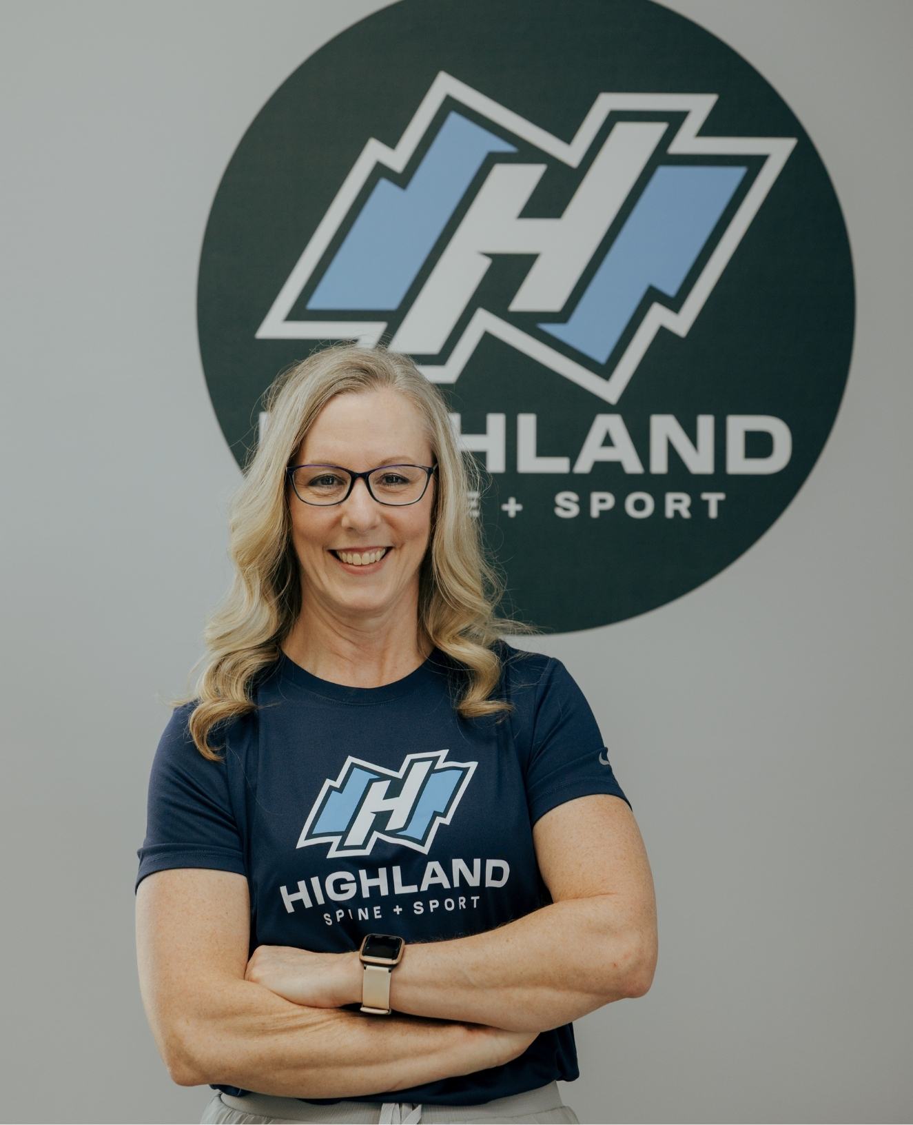 Marie, a middle-aged woman with long, blonde, wavy hair, wearing dark-rimmed glasses and a navy blue t-shirt with the "Highland Spine + Sport" logo, smiles confidently at the camera. She stands against a light-gray wall with a large circular logo featuring a stylized "H" in white and light blue behind her. The image conveys a professional and approachable vibe, emphasizing the brand's identity in sports health and spinal care.