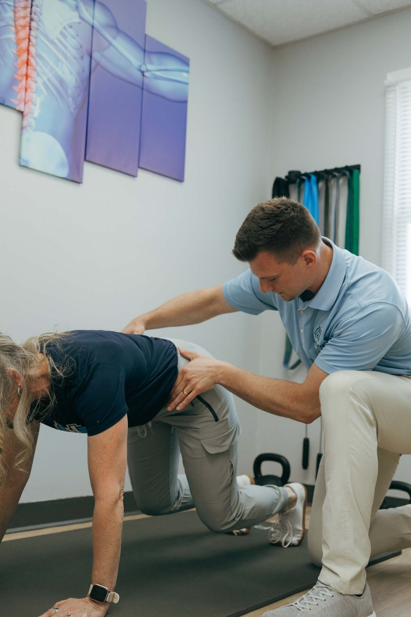Dr. Amos beside a woman on an exercise mat, guiding her form during a rehabilitation exercise to help with hip pain treatment in a bright physical therapy clinic.