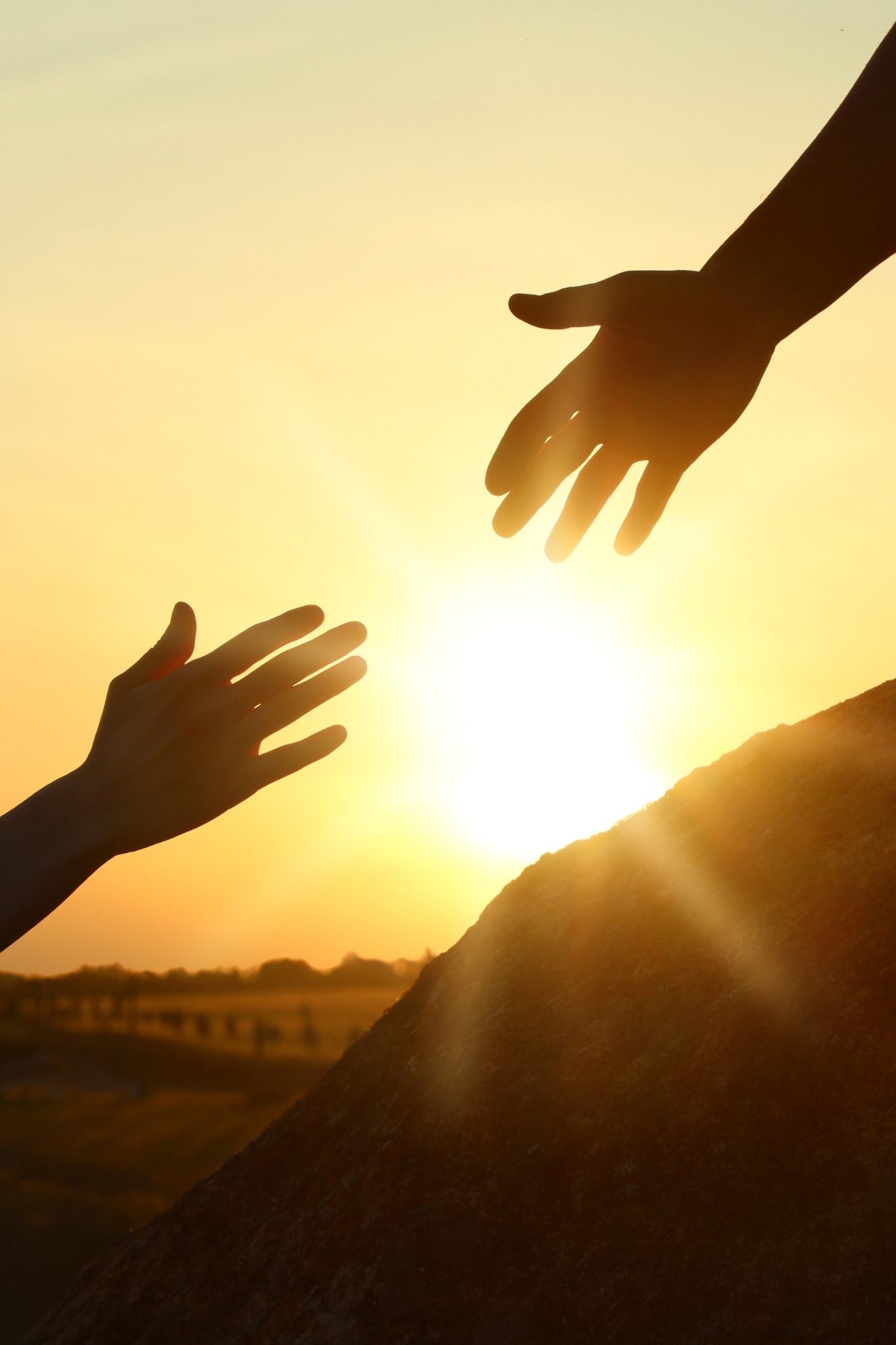 Two silhouetted hands reach toward each other against a bright sunrise, symbolizing support, hope, and helping someone overcome a challenge.