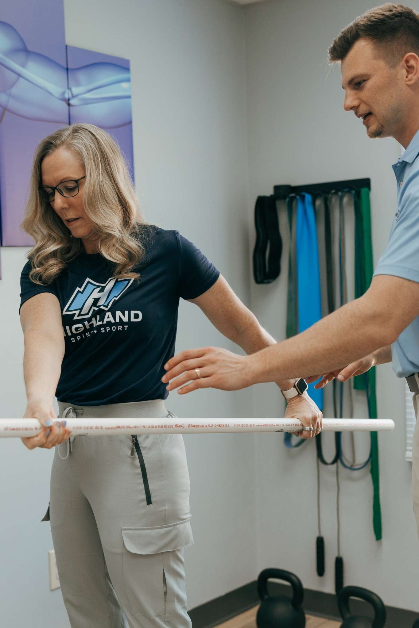 Dr. Amos guides a woman holding a PVC pipe through a shoulder mobility exercise in a physical therapy and sports rehabilitation clinic.