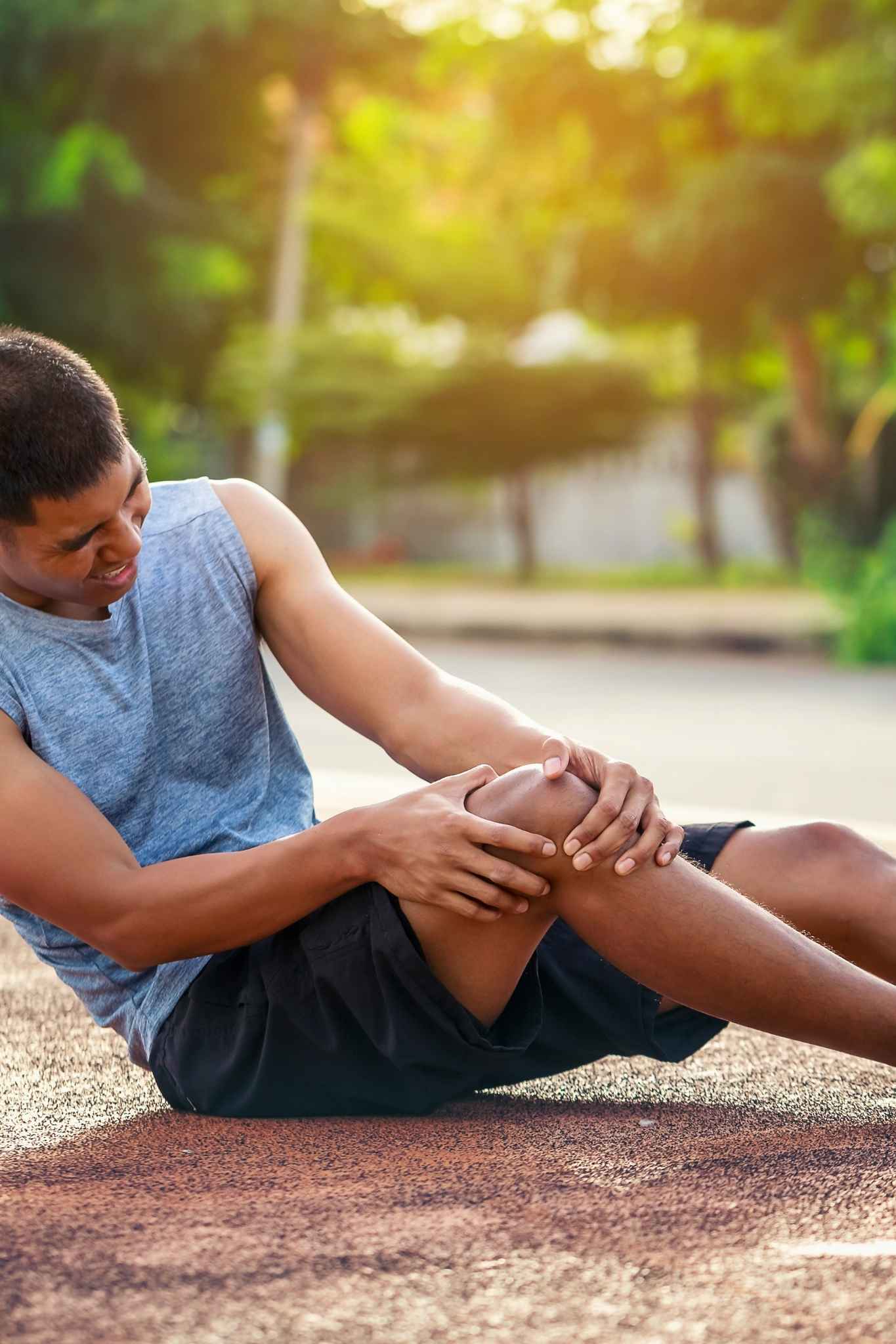 Young male runner sitting on an outdoor track and clutching his right knee in pain, suggesting a sports-related knee injury during exercise.