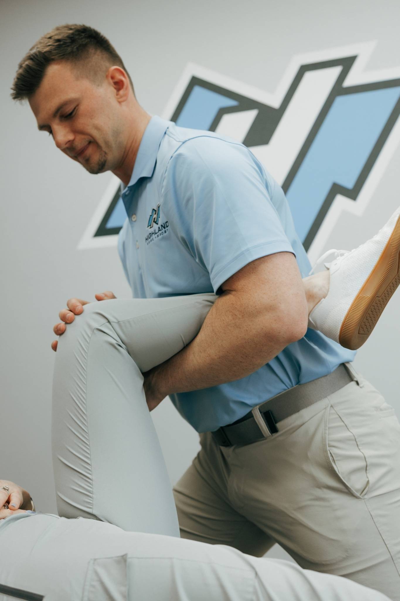 A therapist from Highland Spine + Sport performs a supported leg stretch on a patient lying on a treatment table in a clean clinical setting.