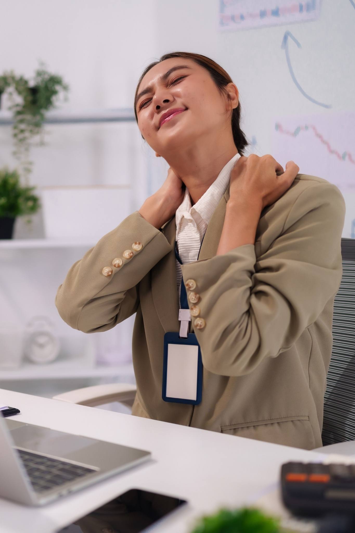 Woman in an office chair holds her neck and shoulders in pain at her desk, suggesting workplace strain or tension.
