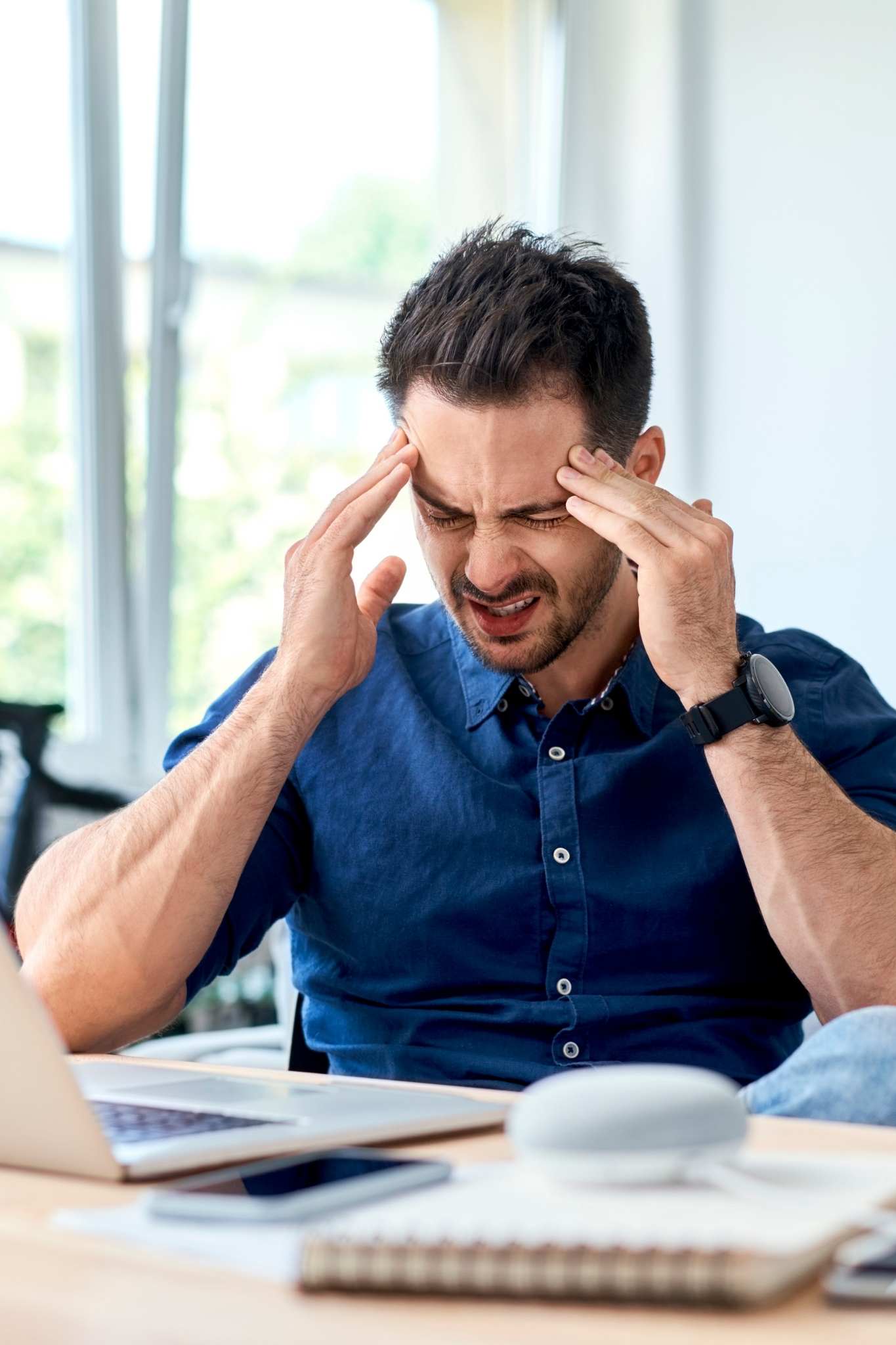 Man in a blue shirt sits at a desk with eyes closed and fingers pressed to his temples, showing stress or headache while working on a laptop.