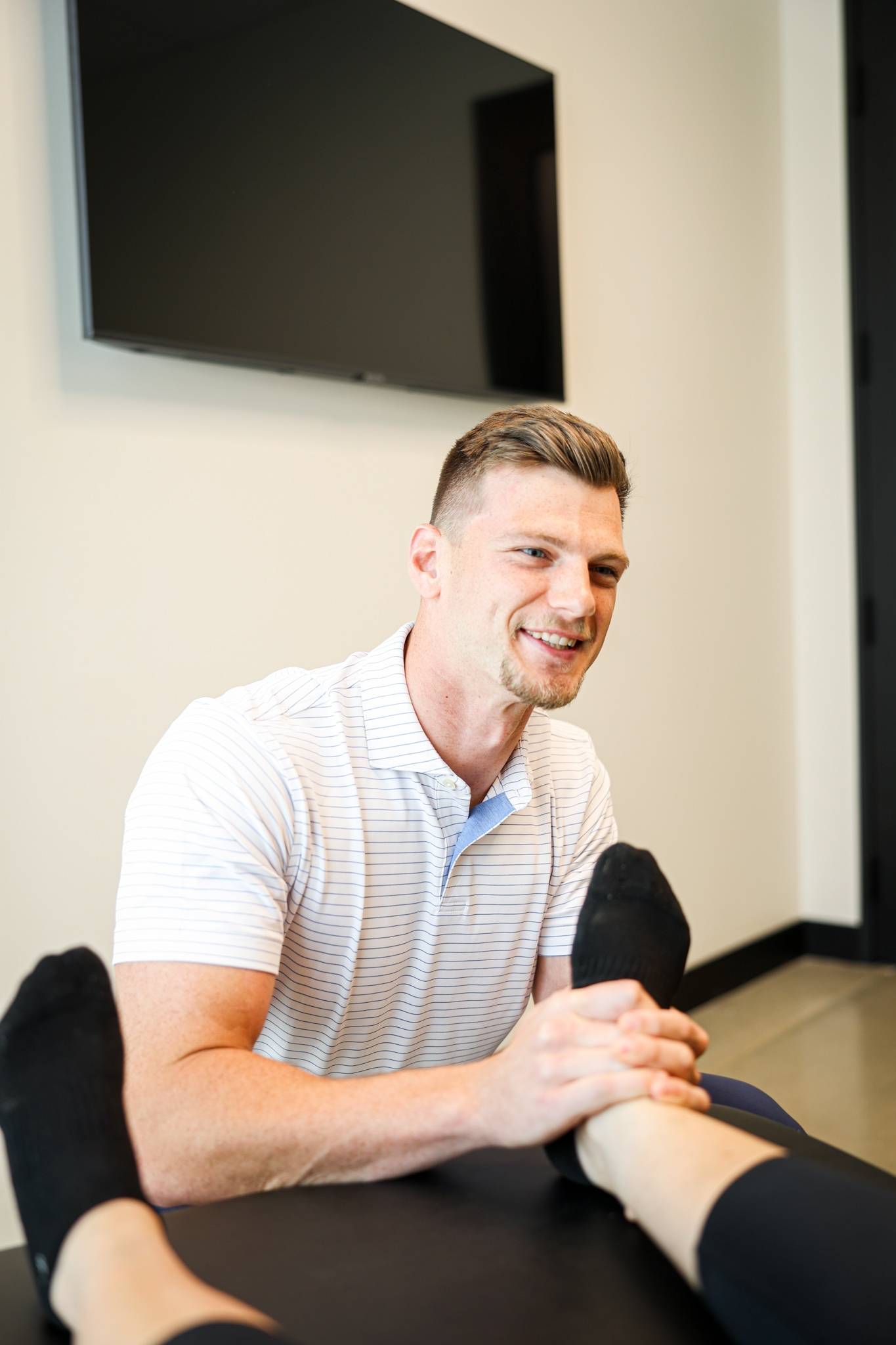 Smiling chiropractor performs soft tissue therapy for ankle or foot mobilization focusing on plantar fasciitis treatment on a patient lying on a treatment table in a bright clinic.
