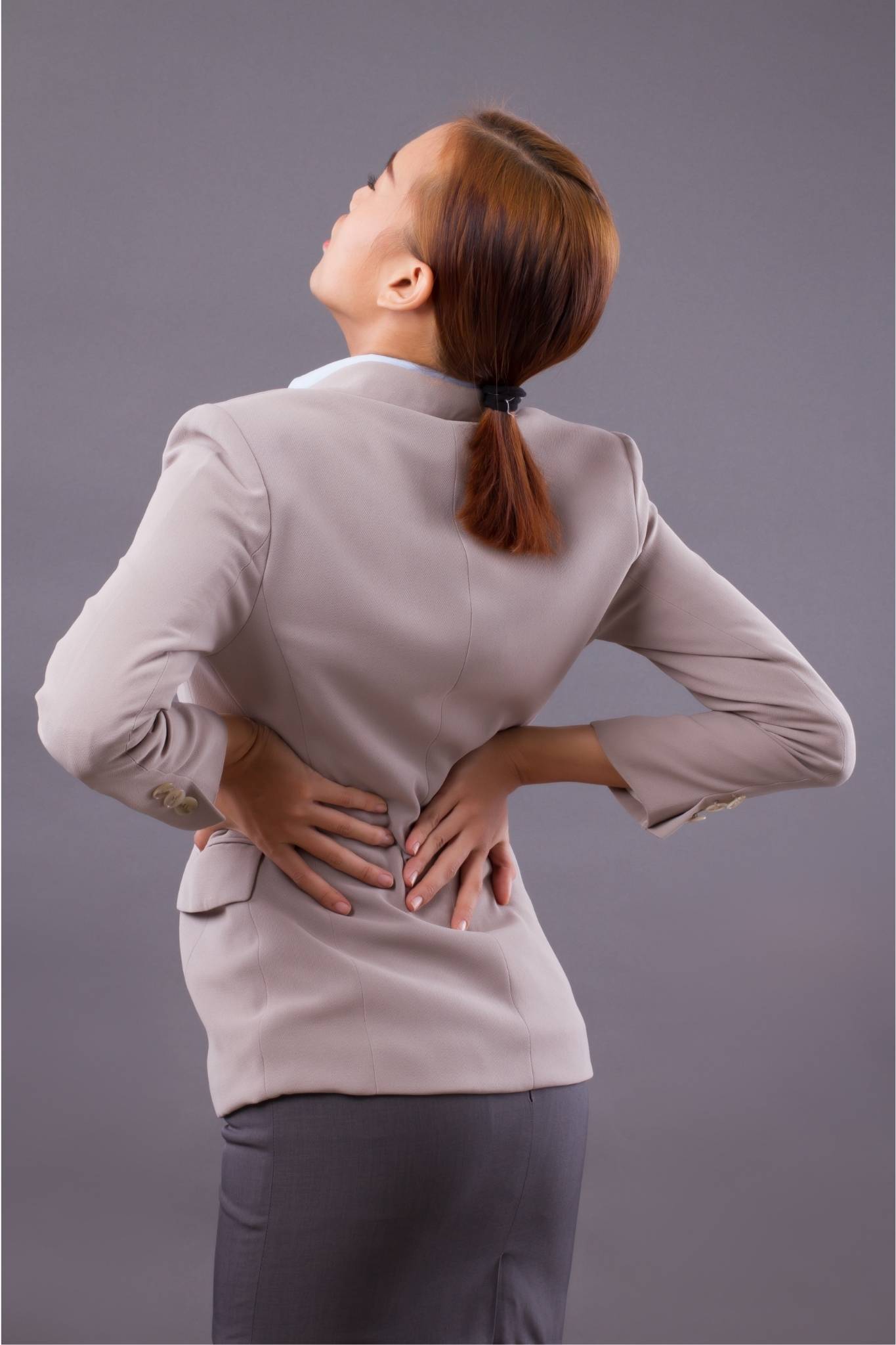 Woman in business attire stands with both hands on her lower back, illustrating back pain, herniated disc, or workplace strain.