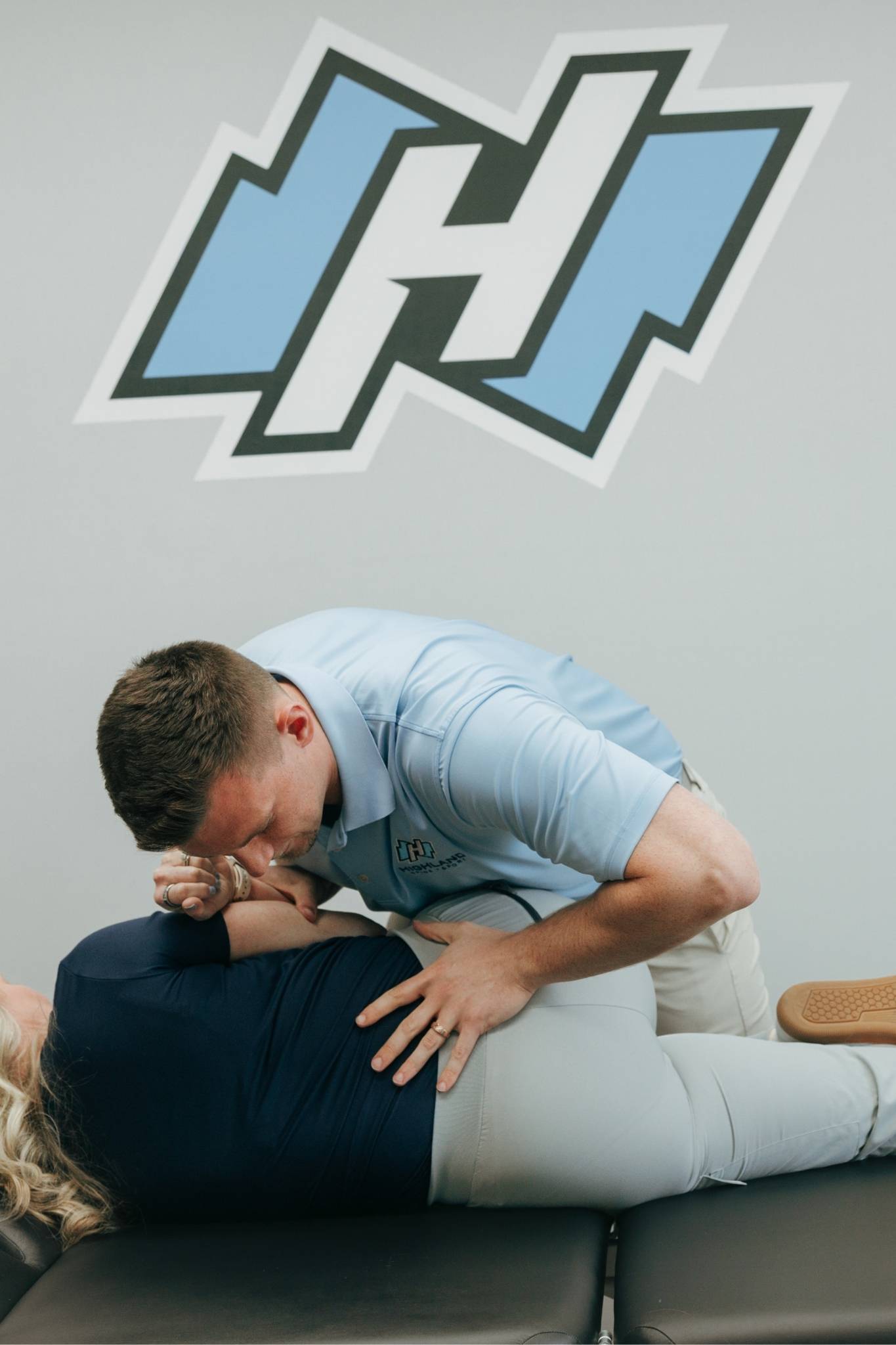 Chiropractor, Dr. Matthew Amos from Highland Spine + Sport, in a blue polo performs a lower back adjustment for a possible herniated disc on a patient lying on a treatment table in a clinic with the light blue and white Highland Spine + Sport logo on the wall.