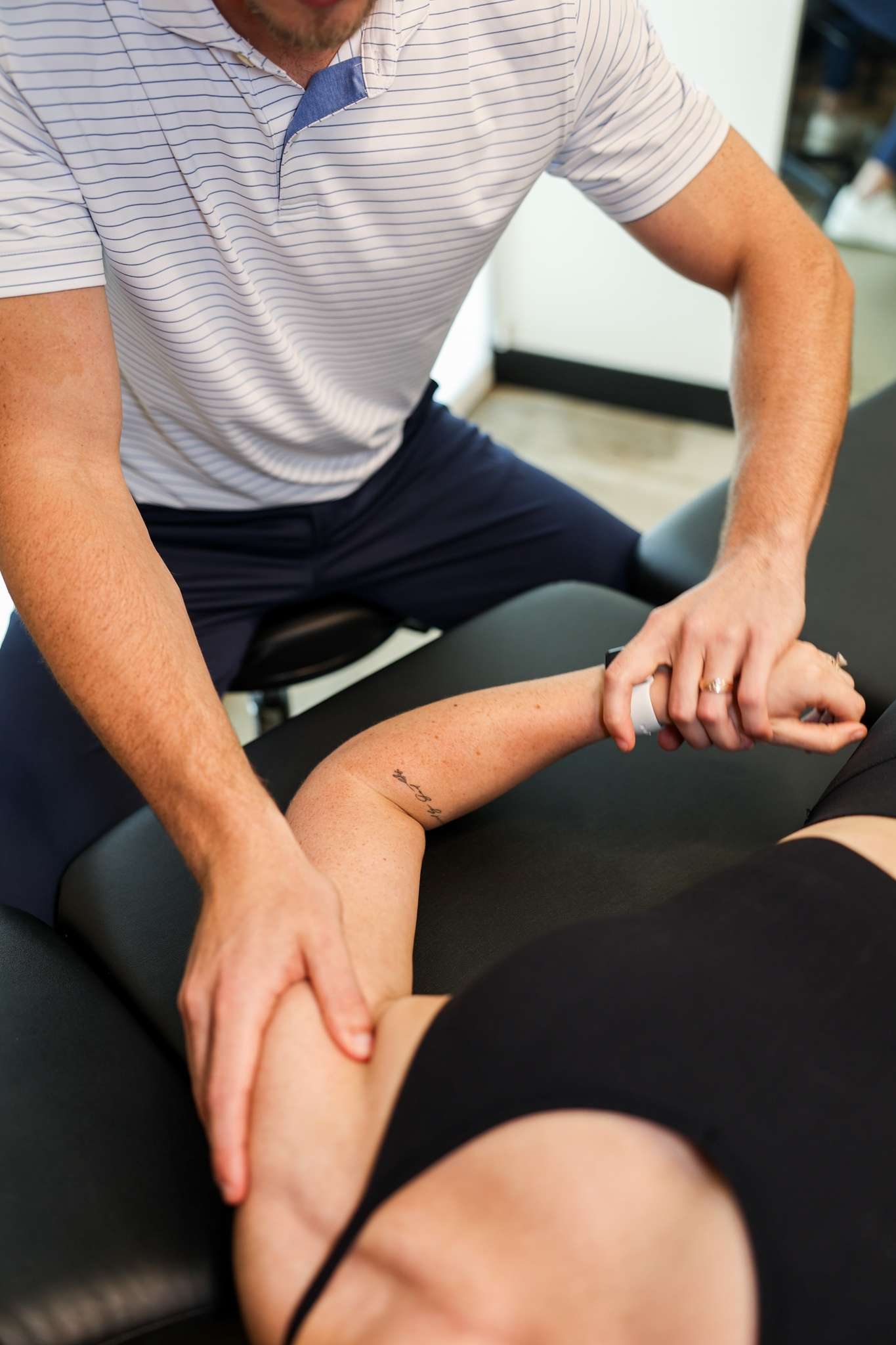 Dr. Matthew Amos performs manual shoulder and elbow therapy on a patient lying on a treatment table in a modern clinic.