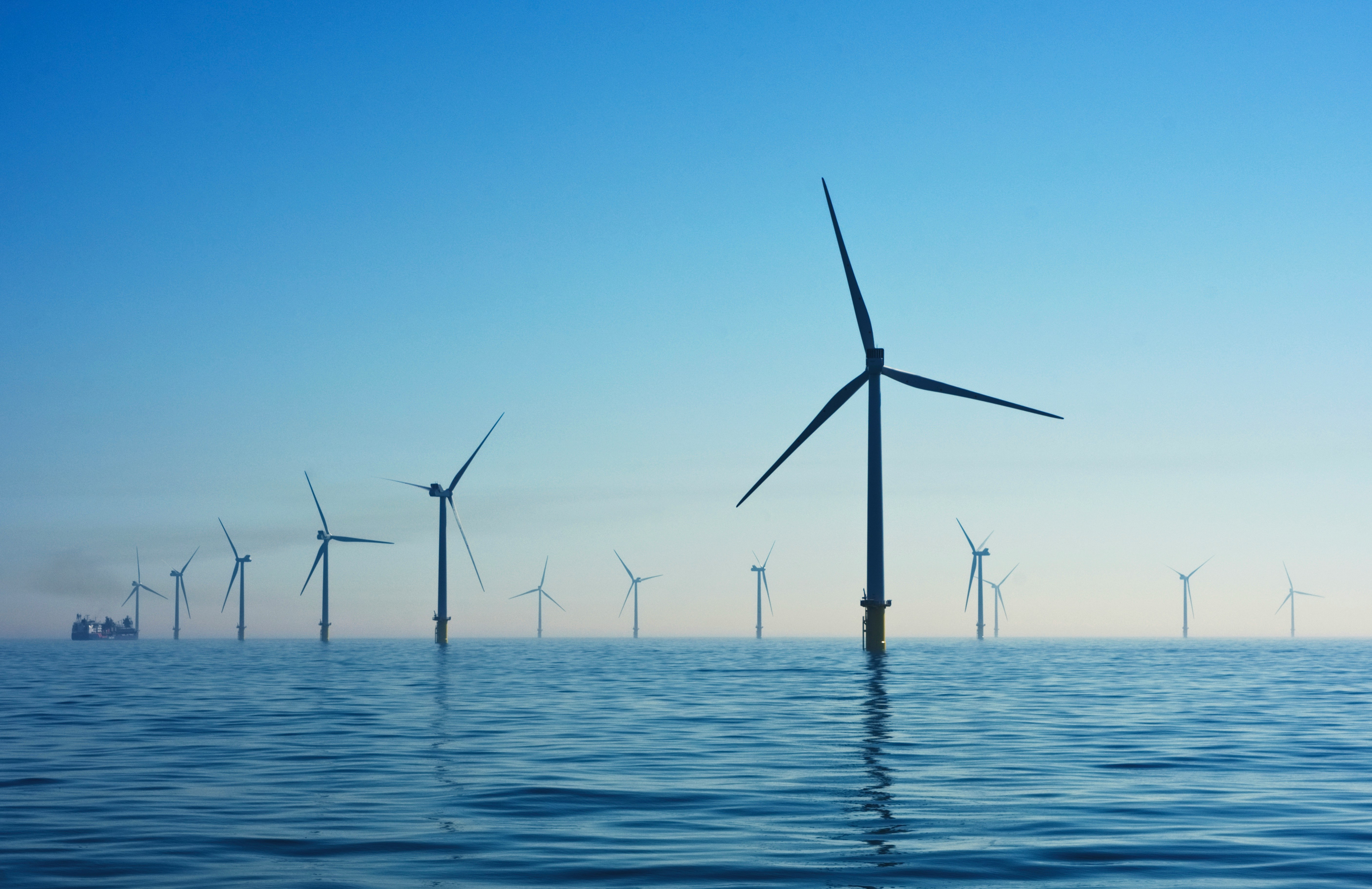 Offshore wind turbines stand in a calm sea under a clear blue sky. A ship can be seen in the distance.