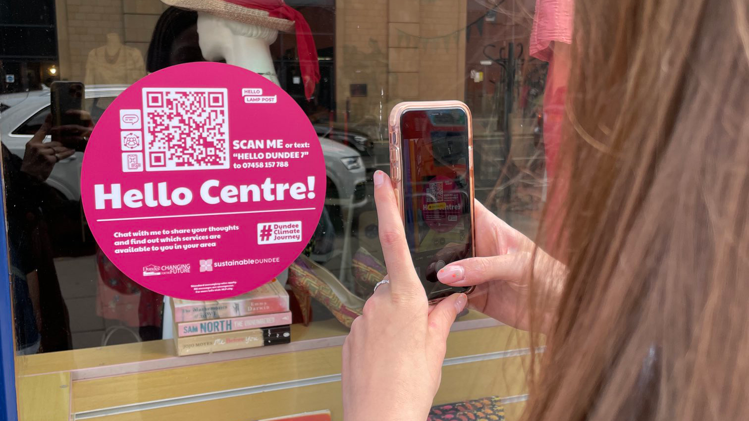 Person using a smartphone to scan a circular pink QR code sticker on a shop window. The window sticker reads "Hello Centre!" with contact information.
