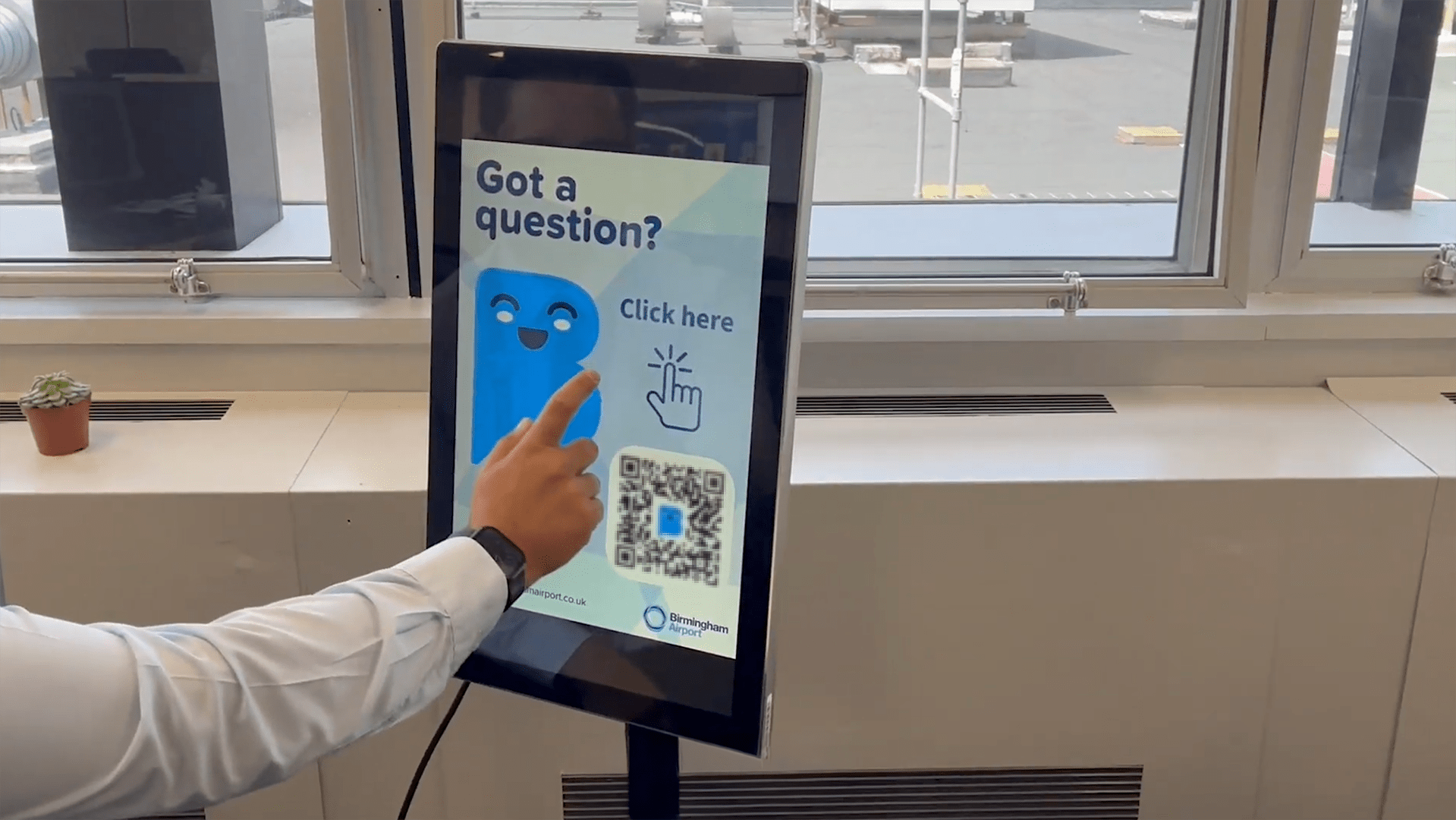 A person in a white shirt interacts with a digital screen displaying "Got a question? Click here" and a QR code.