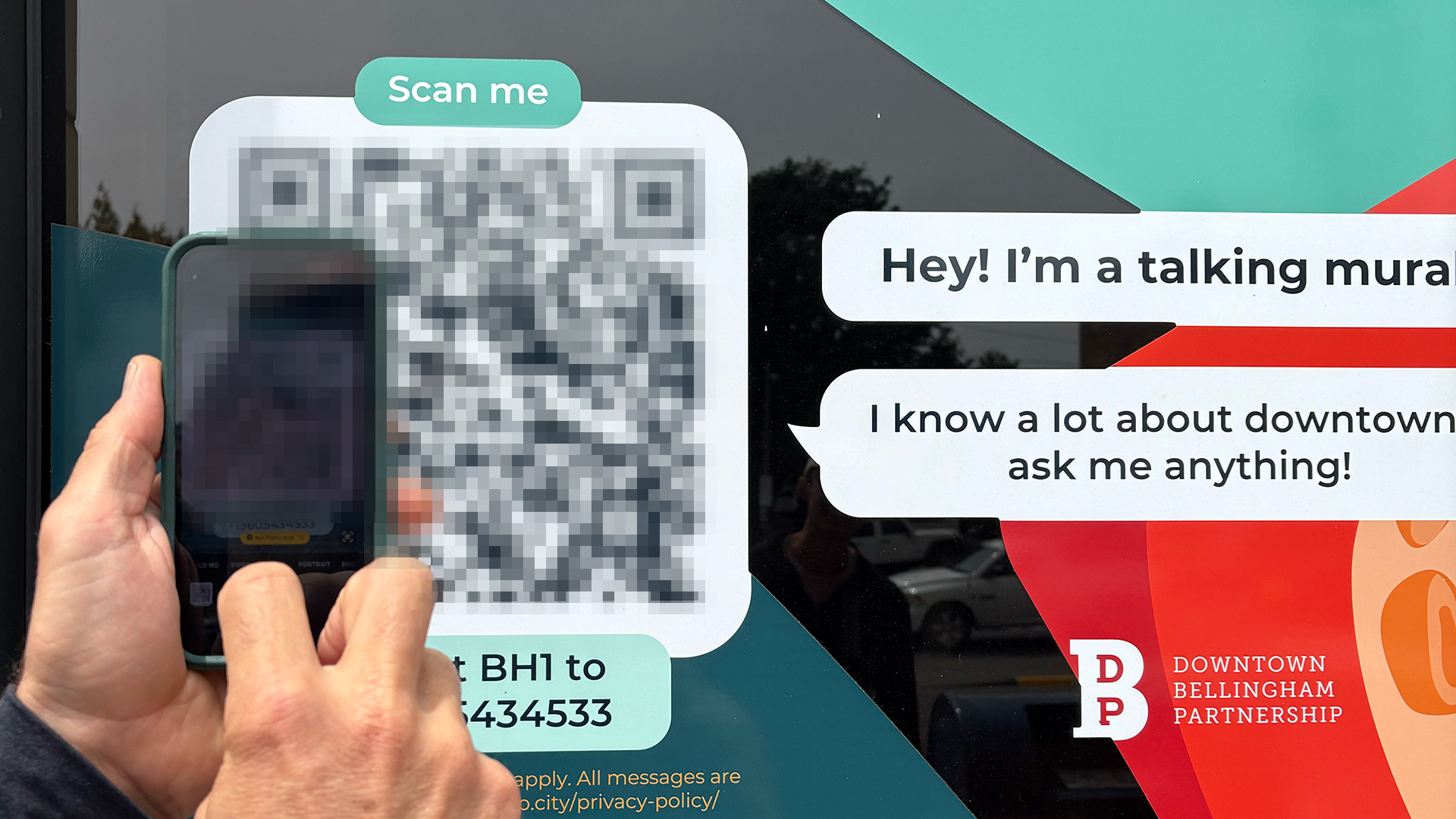 A person scans a large QR code on a window with a smartphone. Text bubbles say, "Hey! I'm a talking mural," promoting downtown Bellingham