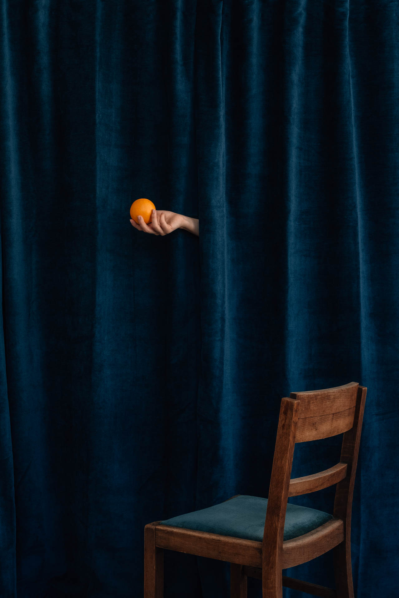 The hand of Saint Citrus holding an orange in front of blue velvet above an empty chair