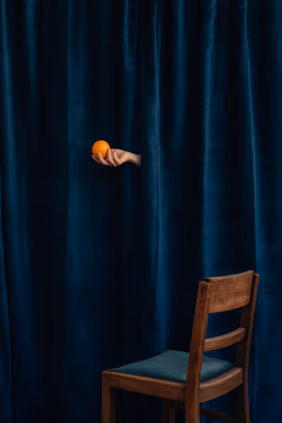 The hand of Saint Citrus holding an orange in front of blue velvet above an empty chair