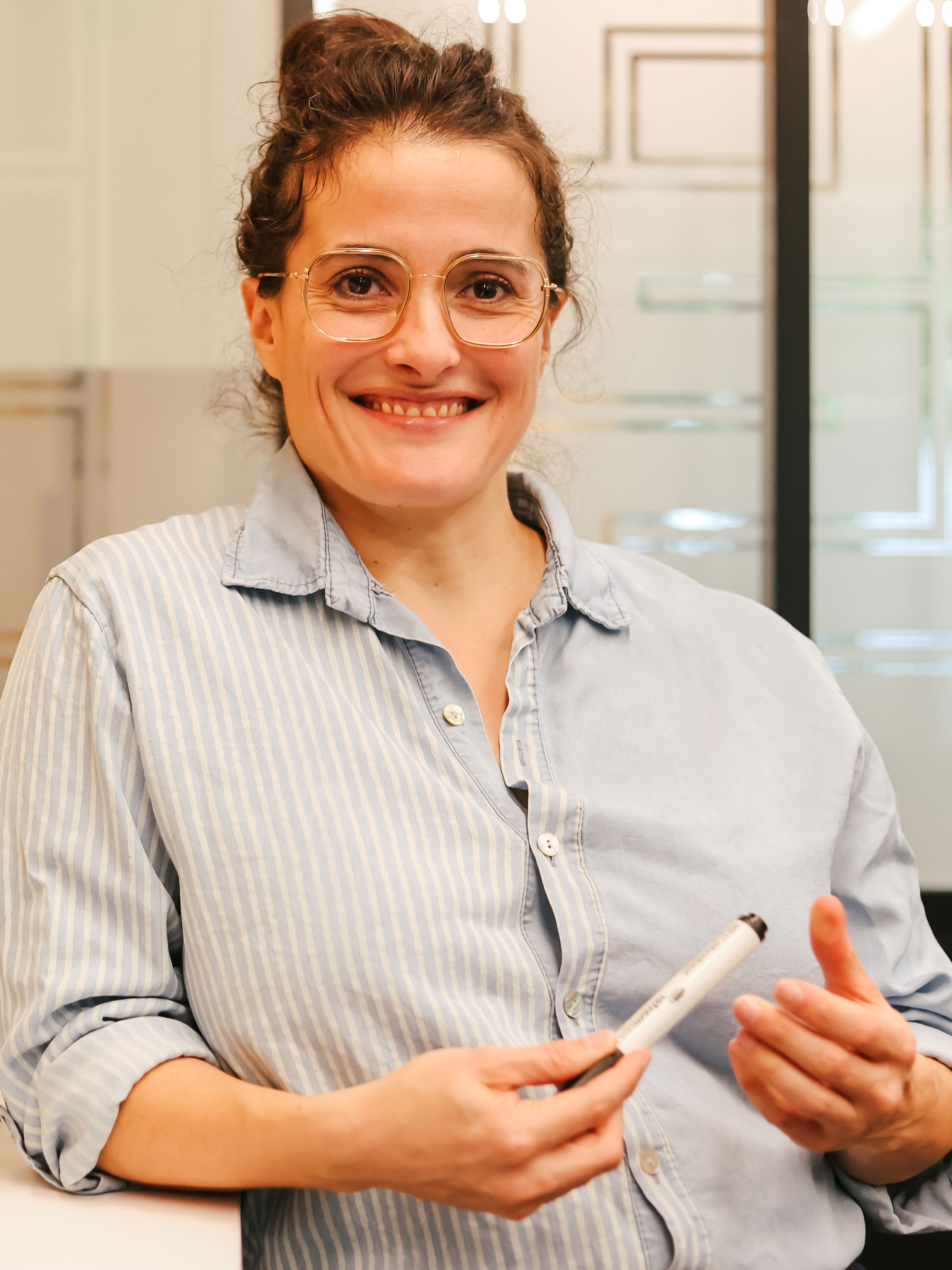Femme souriante portant des lunettes et une chemise à rayures, tenant un marqueur.