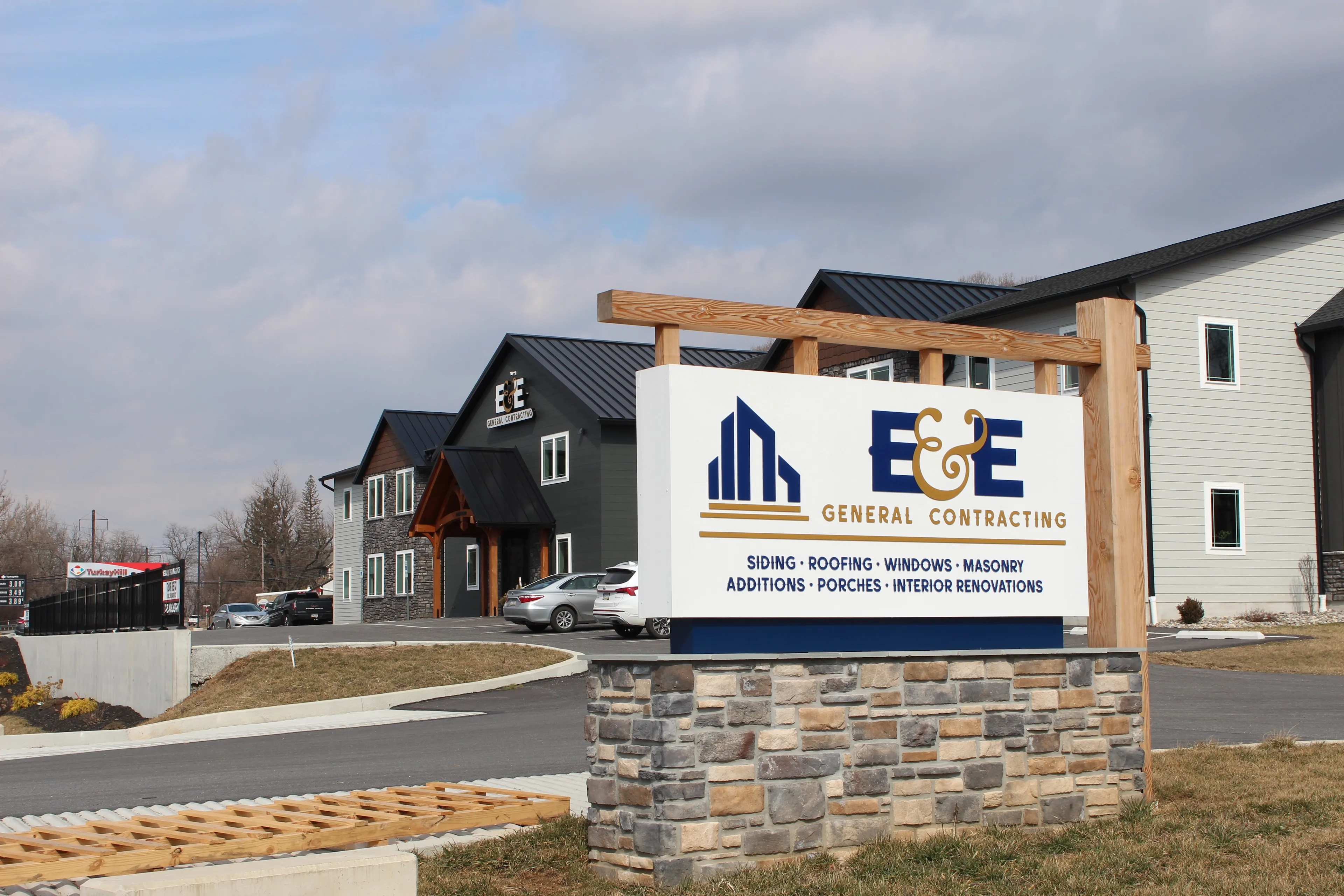 A large sign for E&E General Contracting stands in front of a modern office building with cars parked outside.