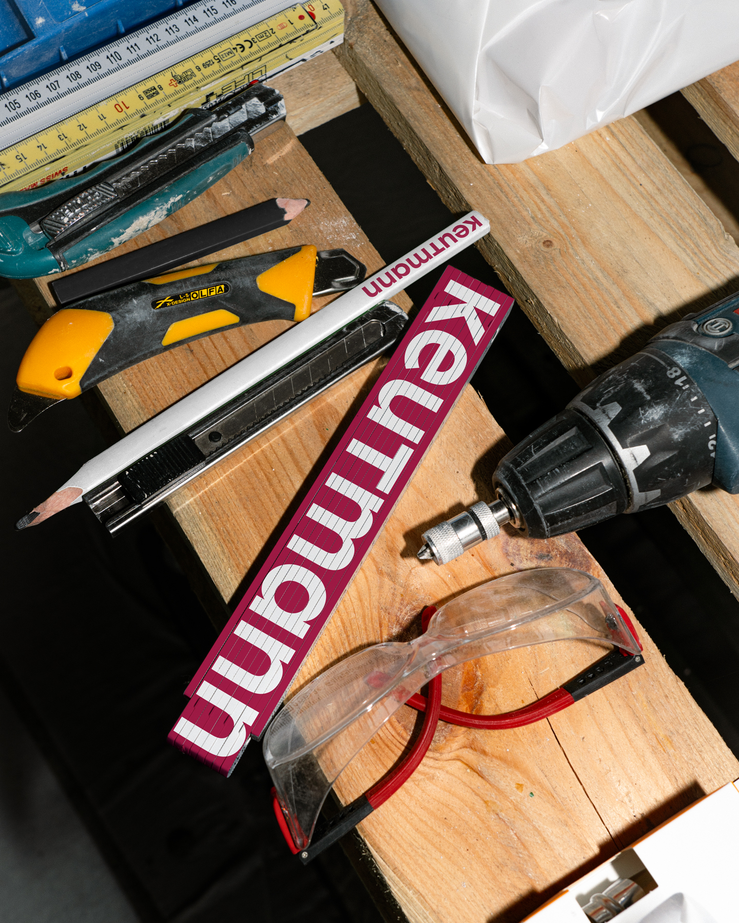 Werkzeuge auf Holzbrett, darunter Schutzbrille, Bohrmaschine, Lineal und zwei Cutter-Messer.