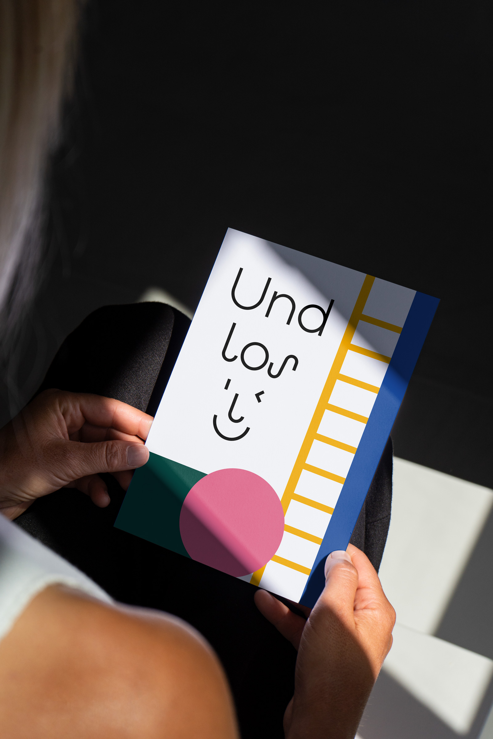 Person hält eine farbenfrohe Karte mit abstraktem Gesicht und Schriftzug ‚Und los‘.