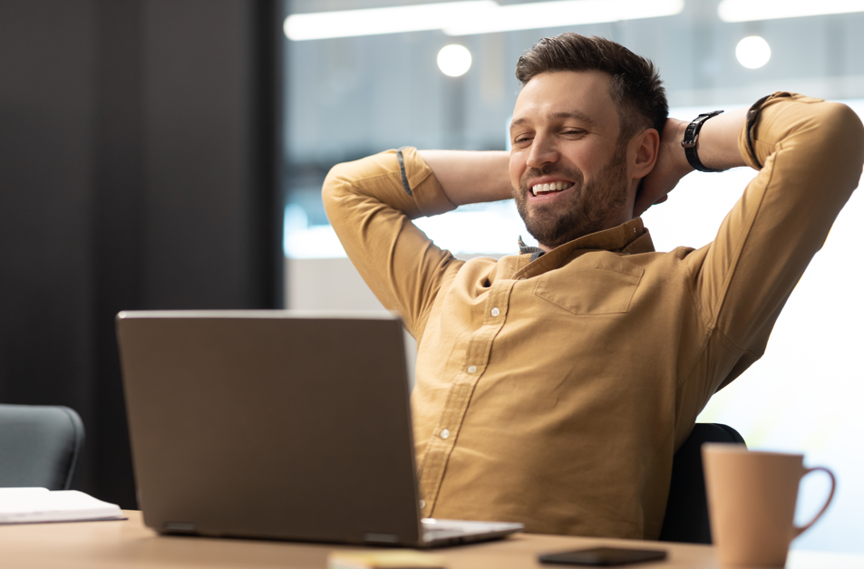 Happy Client. A smiling man in a mustard shirt leans back, hands behind his head, in front of a laptop at a desk.