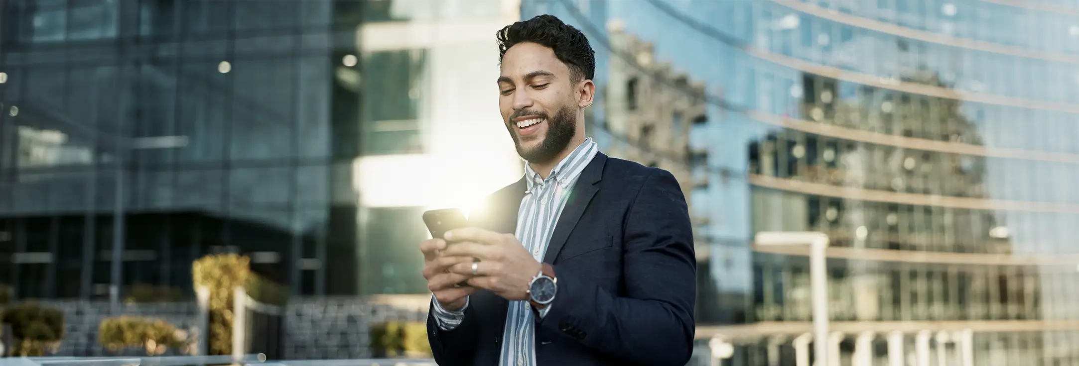 Smiling man in a blazer standing outdoors, looking at his smartphone with sunlight reflecting on the screen.