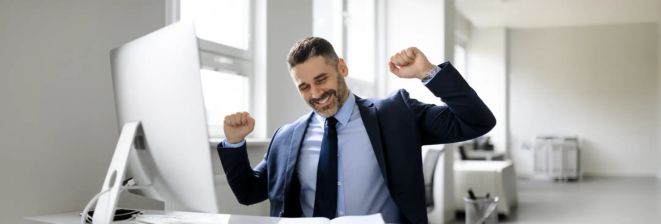 Smiling businessman in suit stretching with raised arms at his office desk.