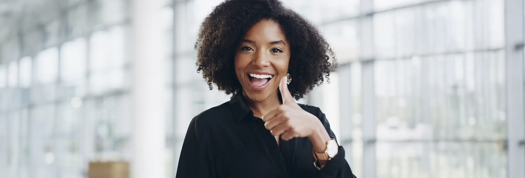 Smiling woman with curly hair wearing a black shirt showing a thumbs-up gesture indoors.
