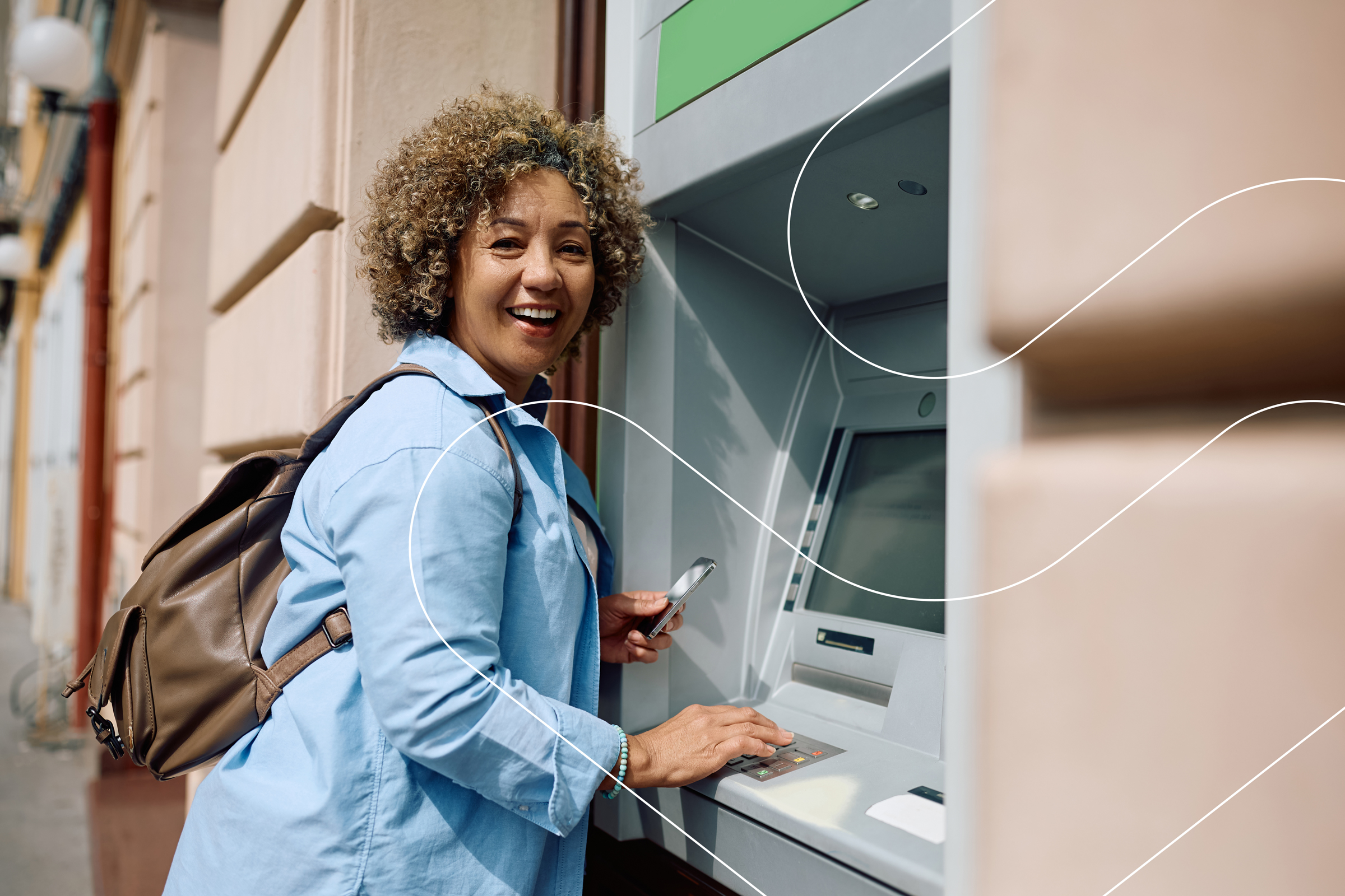 Smiling woman with backpack using ATM outdoors, holding phone in one hand and pressing keypad.