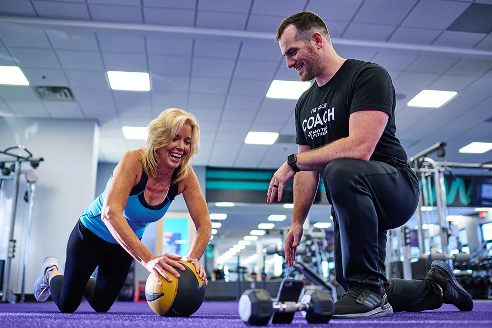 A personal trainer supervising a client doing an exercise with a medicine ball in a gym.