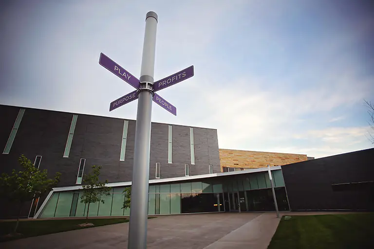Signpost with "Play, Profits, Purpose, People" in front of modern building.