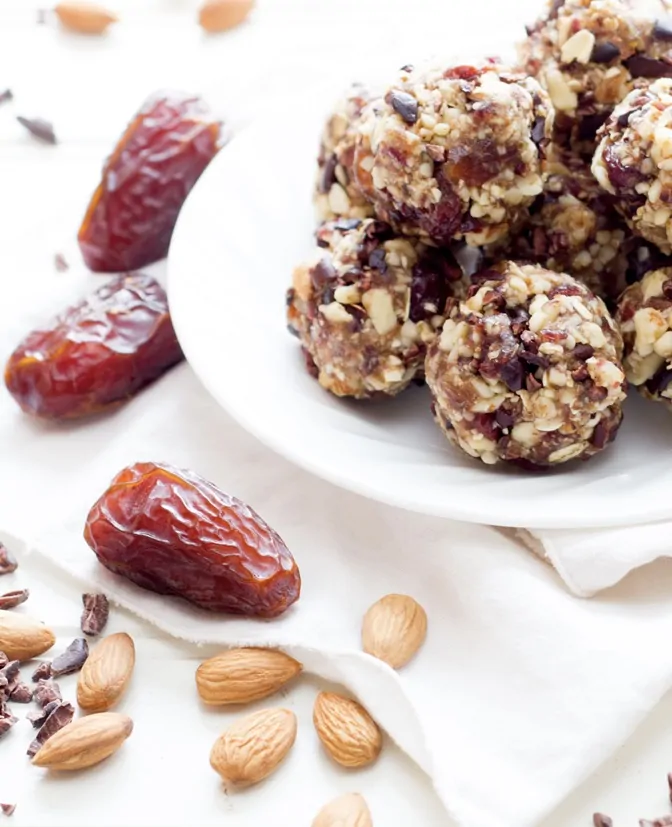 A plate of energy balls made from dates, nuts, and cacao next to whole dates and almonds on a light background.