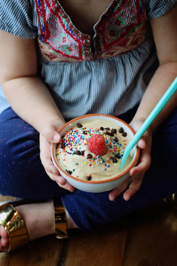 A child holds a bowl of ice cream topped with chocolate chips, colorful sprinkles, and a raspberry, with a bright teal spoon.