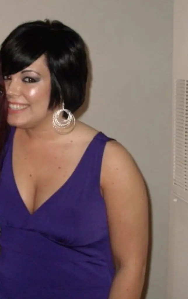 A person wearing a sleeveless purple dress and large silver hoop earrings, standing against a neutral wall background.