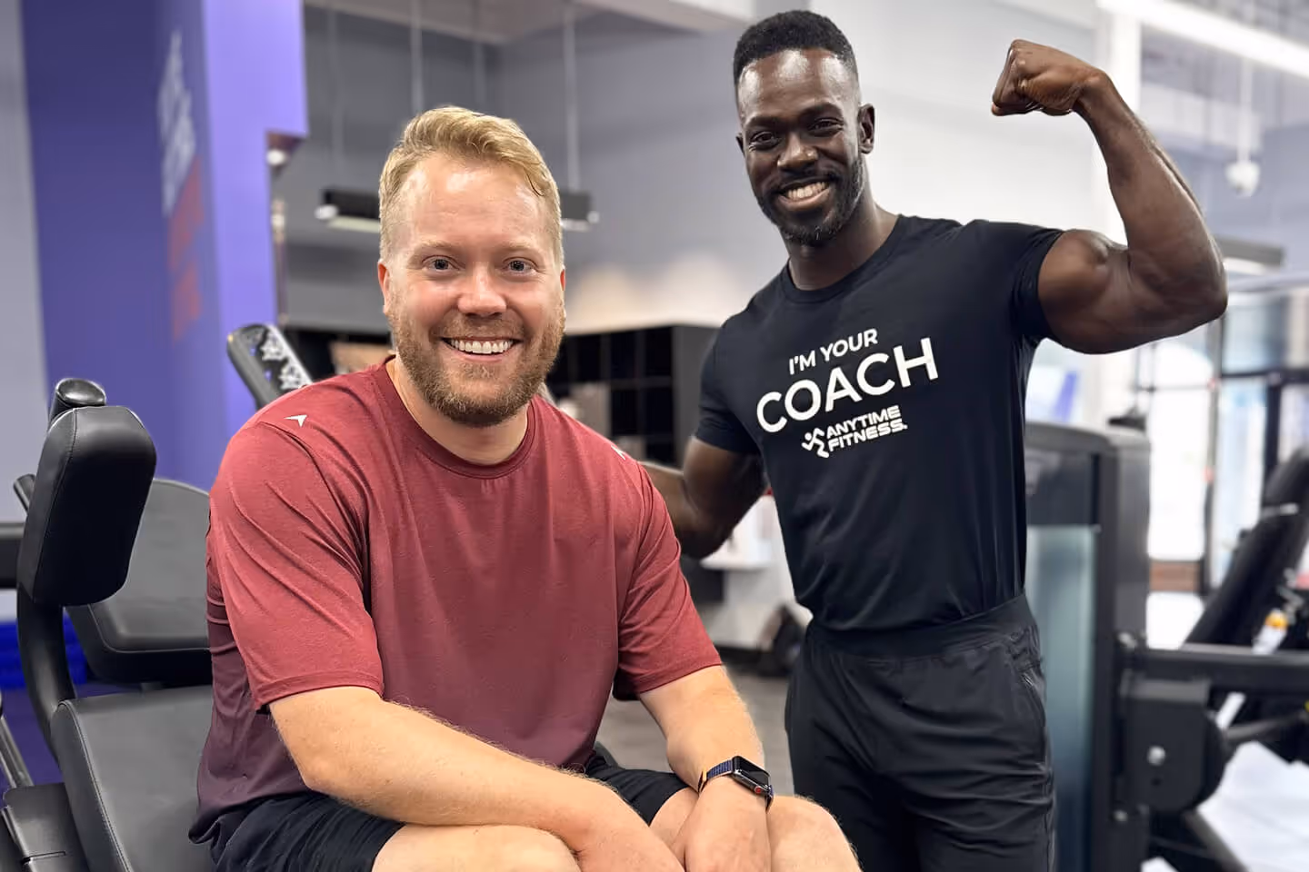 Two smiling men at a gym, one sitting and one flexing his arm, wearing an 'I'm your coach' shirt