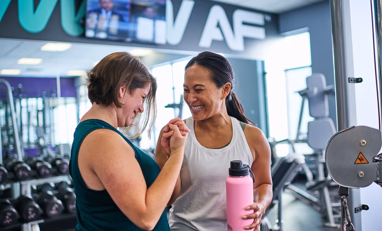 Two women smiling and giving a fist bump at the gym, holding a water bottle after a workout.