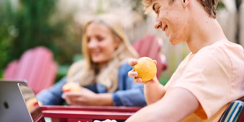 Smiling young man eating an apple while working on a laptop outdoors with a woman in the background.