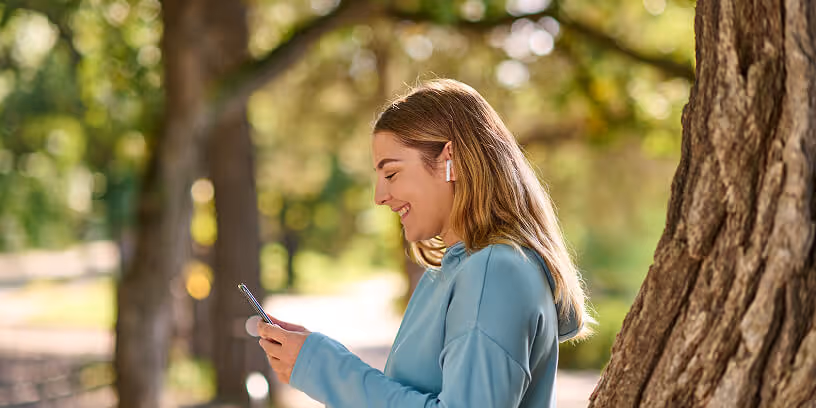 Woman smiling while using her phone and wearing earbuds outdoors near a tree.