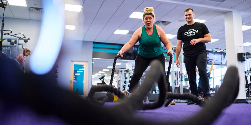 Woman working out with battle ropes at the gym while a coach looks on.