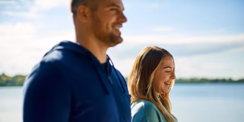 Smiling man and woman walking outdoors near the water on a sunny day.
