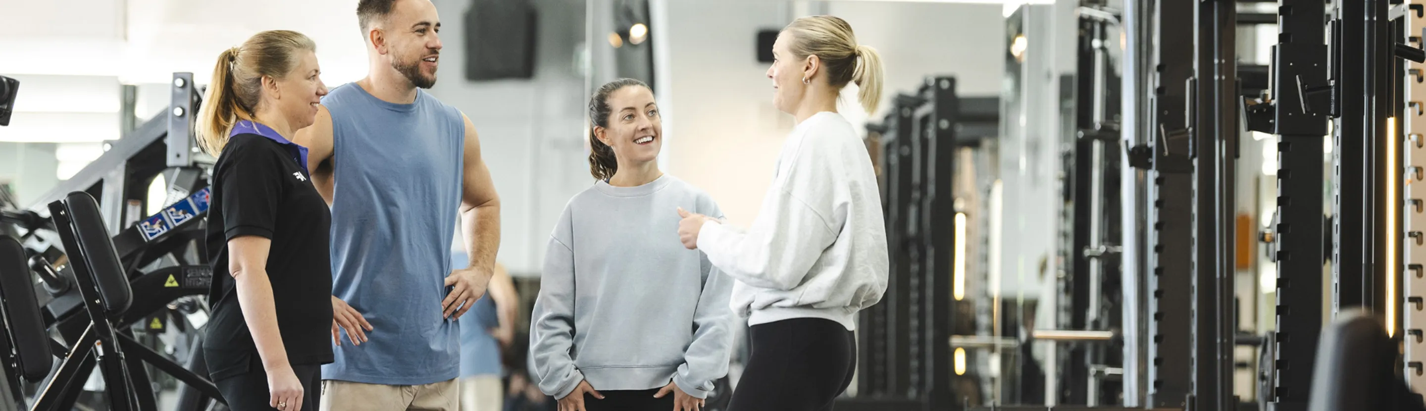 A group of four individuals converses in a gym, surrounded by exercise equipment, discussing fitness routines and training.