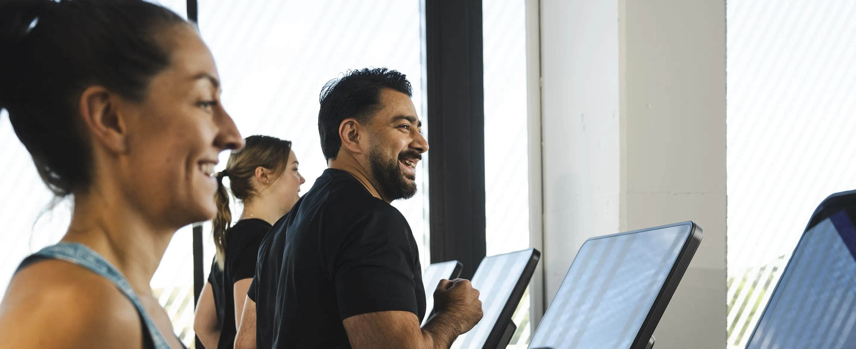 A group of individuals workout together on treadmills, focused and engaged against a backdrop of large windows and natural light
