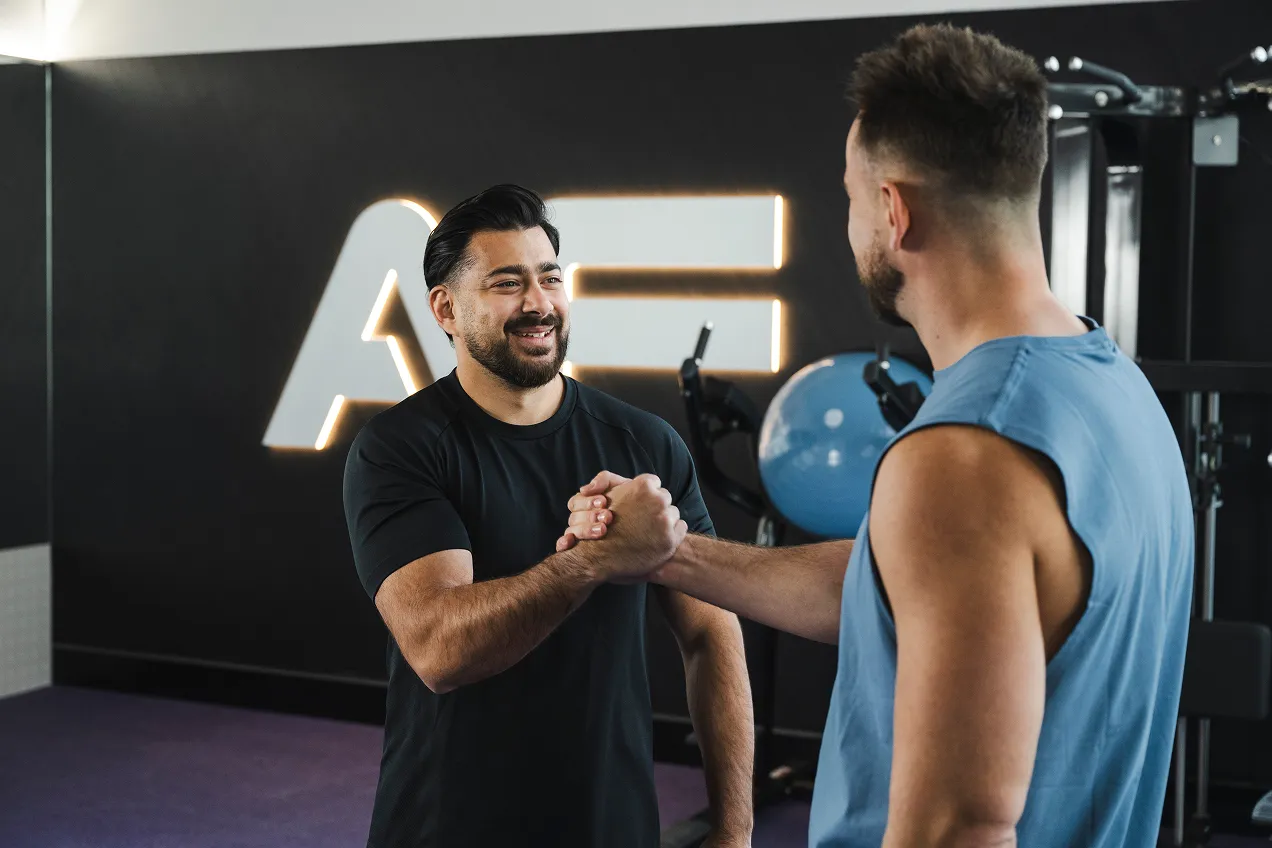 Two men greet each other with a handshake in a modern gym, featuring sleek equipment and a stylish logo in the background.