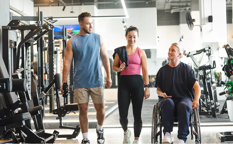 Three individuals in gym attire walking together, with fitness equipment visible in the background, promoting an inclusive fitness environment.