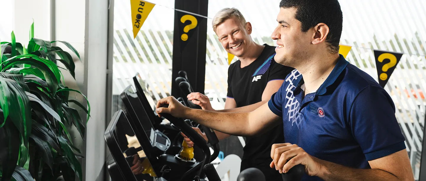 Two men engage in a fitness activity featuring exercise machines, surrounded by lush green plants and cheerful promotional banners.